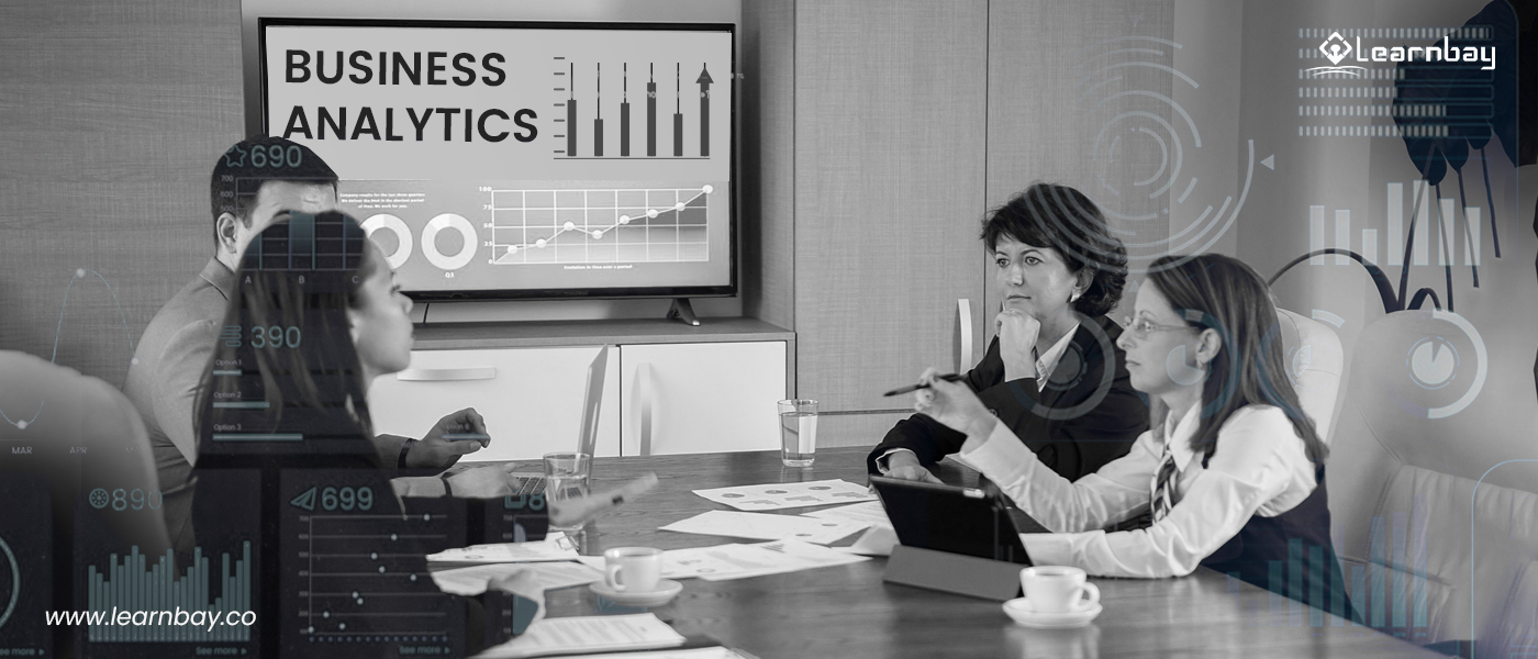 A photo shows a group of business analysts discussing on a project with graphical presentation. The LED screen in the room presents different graphs like, 'Stock charts', 'Area charts' and 'donut charts'.